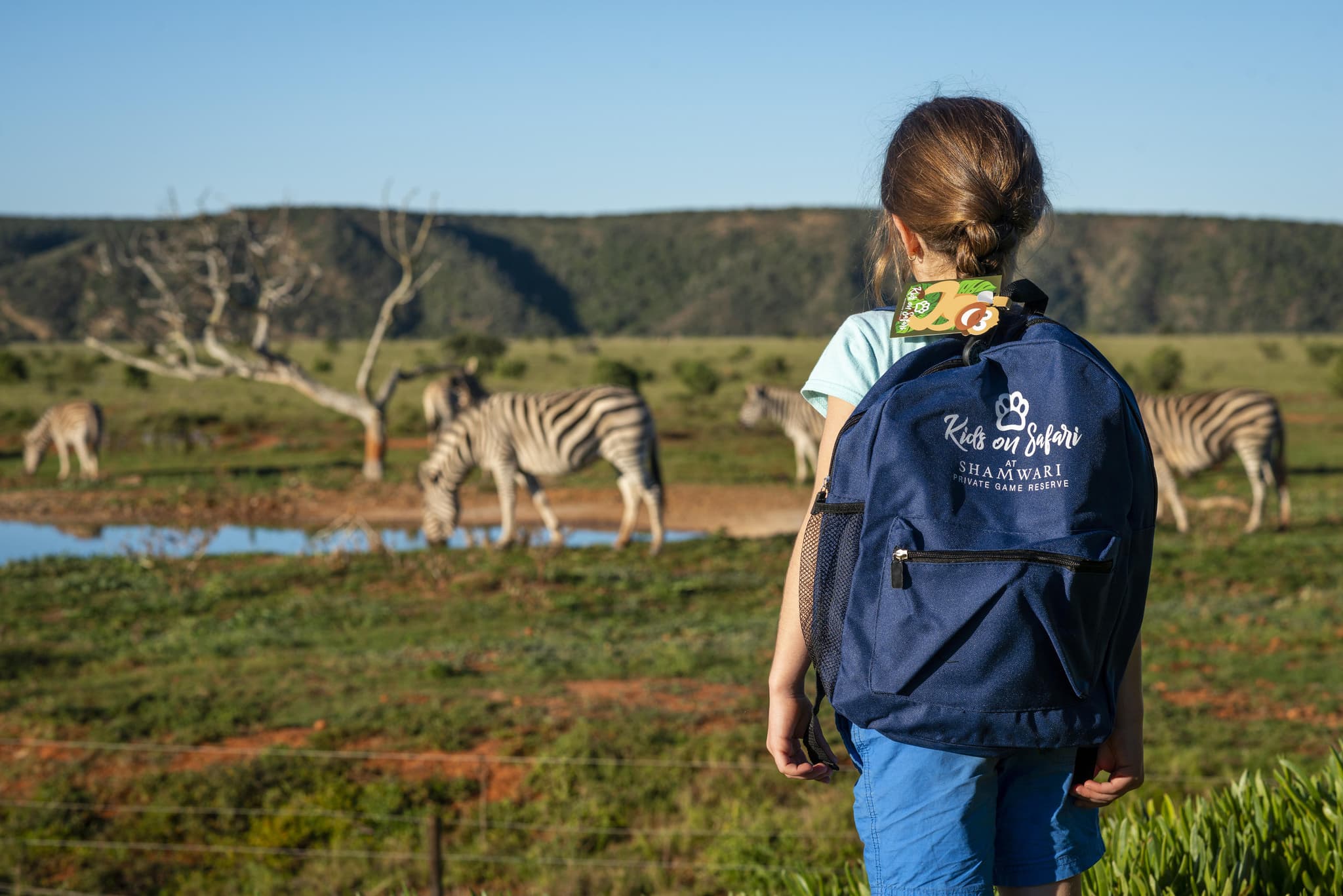 Young girl with Shamwari Kids on Safari backpack watching zebras at a waterhole