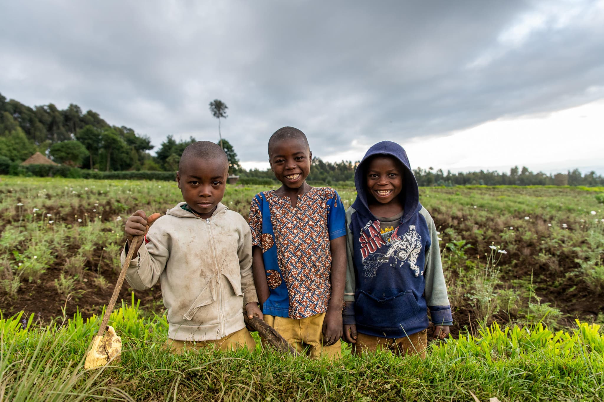 Three smiling children in a lush green field under dramatic skies