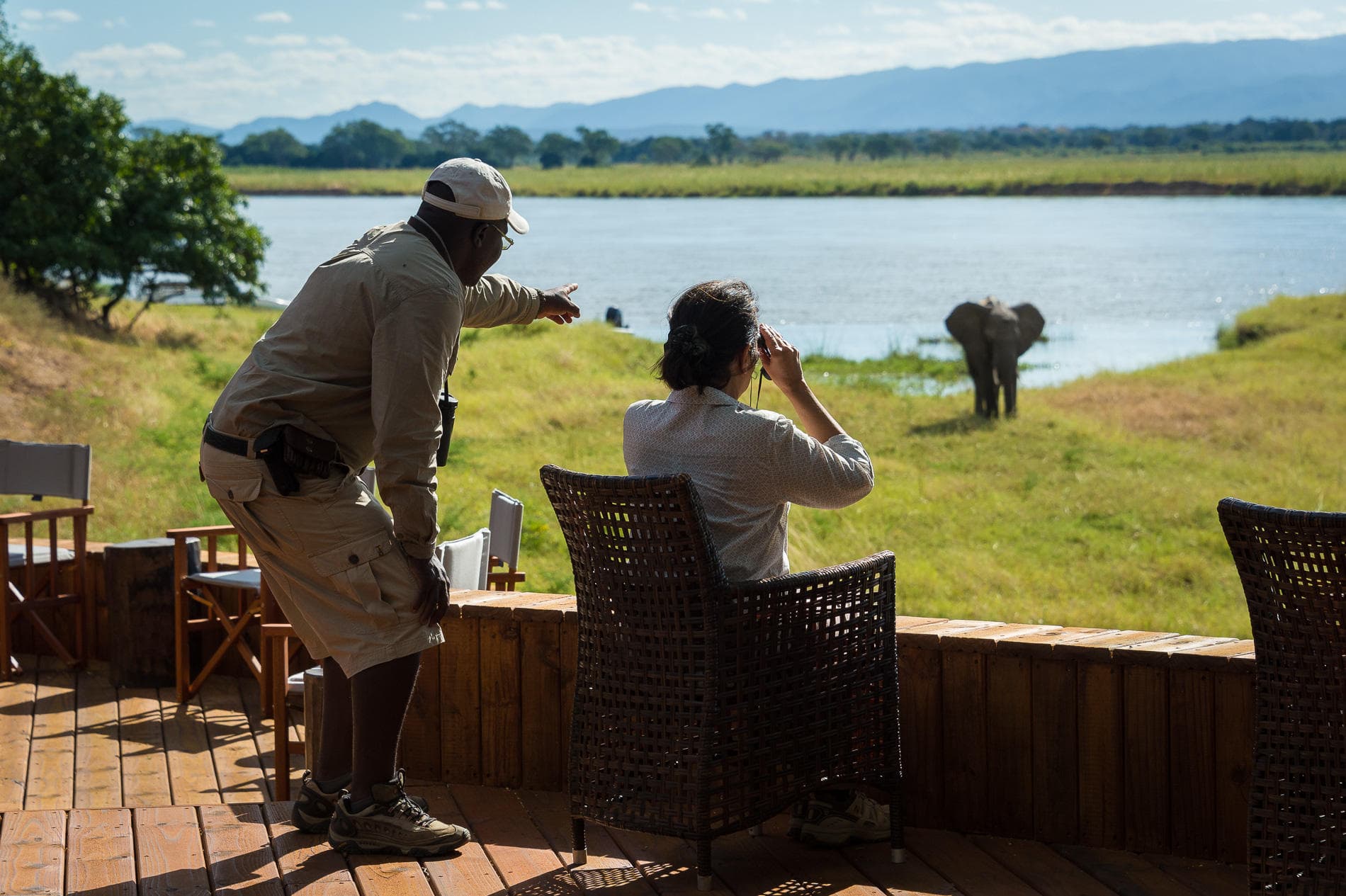 Guide and guest with binoculars watching an elephant crossing a river from a lodge deck