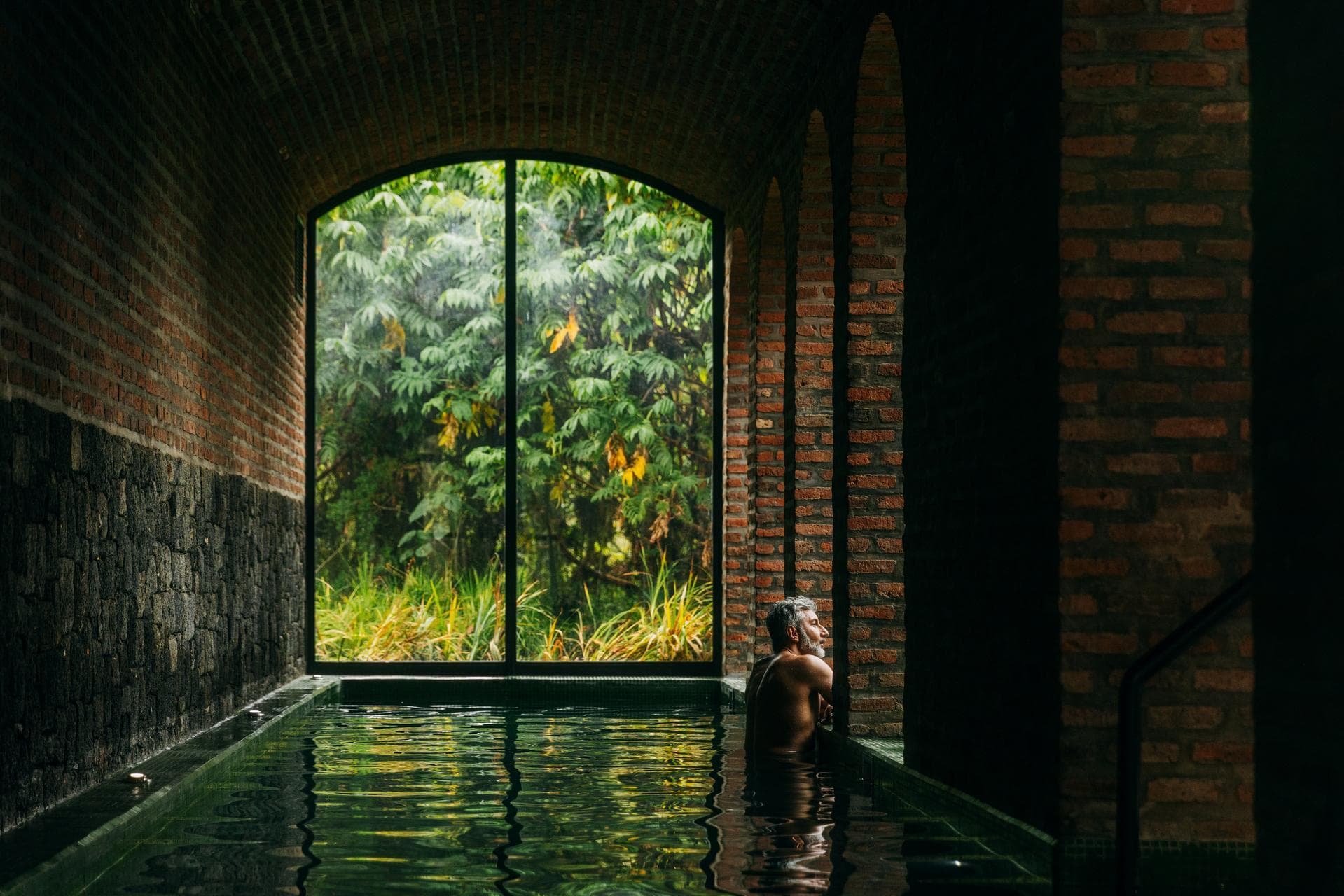 Man relaxing in a brick-vaulted spa pool looking out through an arched window at lush green foliage
