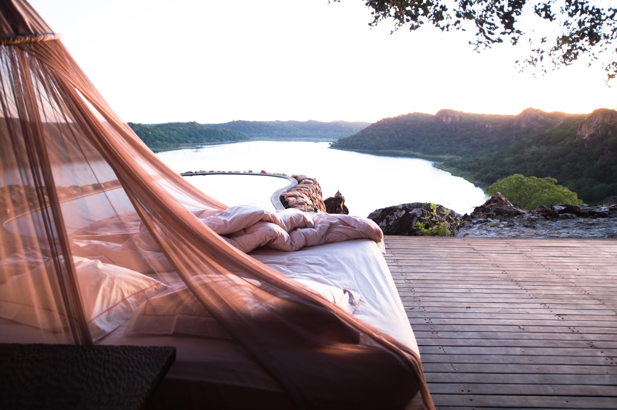 Star bed with flowing mosquito net on a wooden deck overlooking a river winding through hills at sunset
