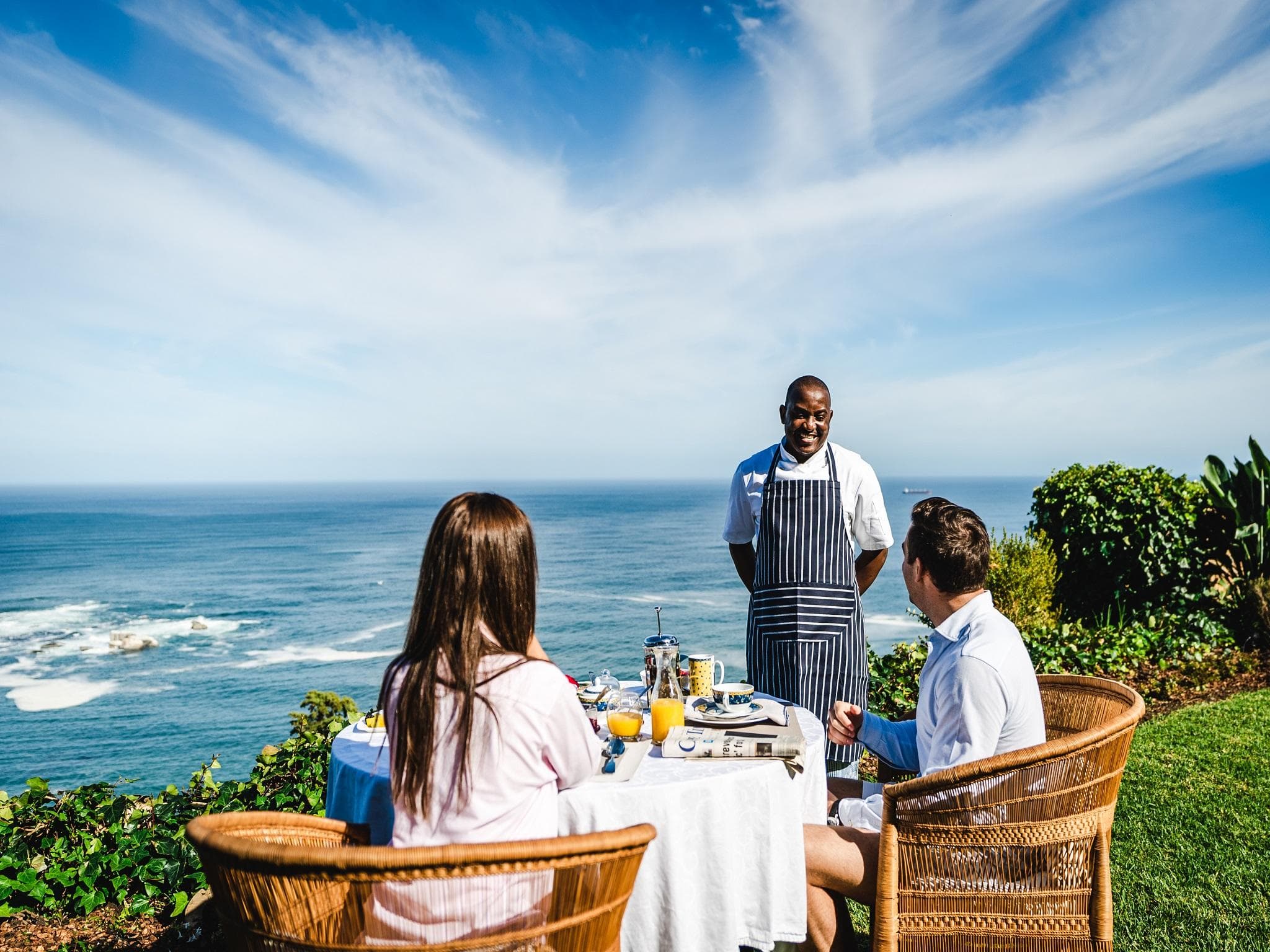 Couple enjoying breakfast served by a chef at a clifftop table overlooking the ocean