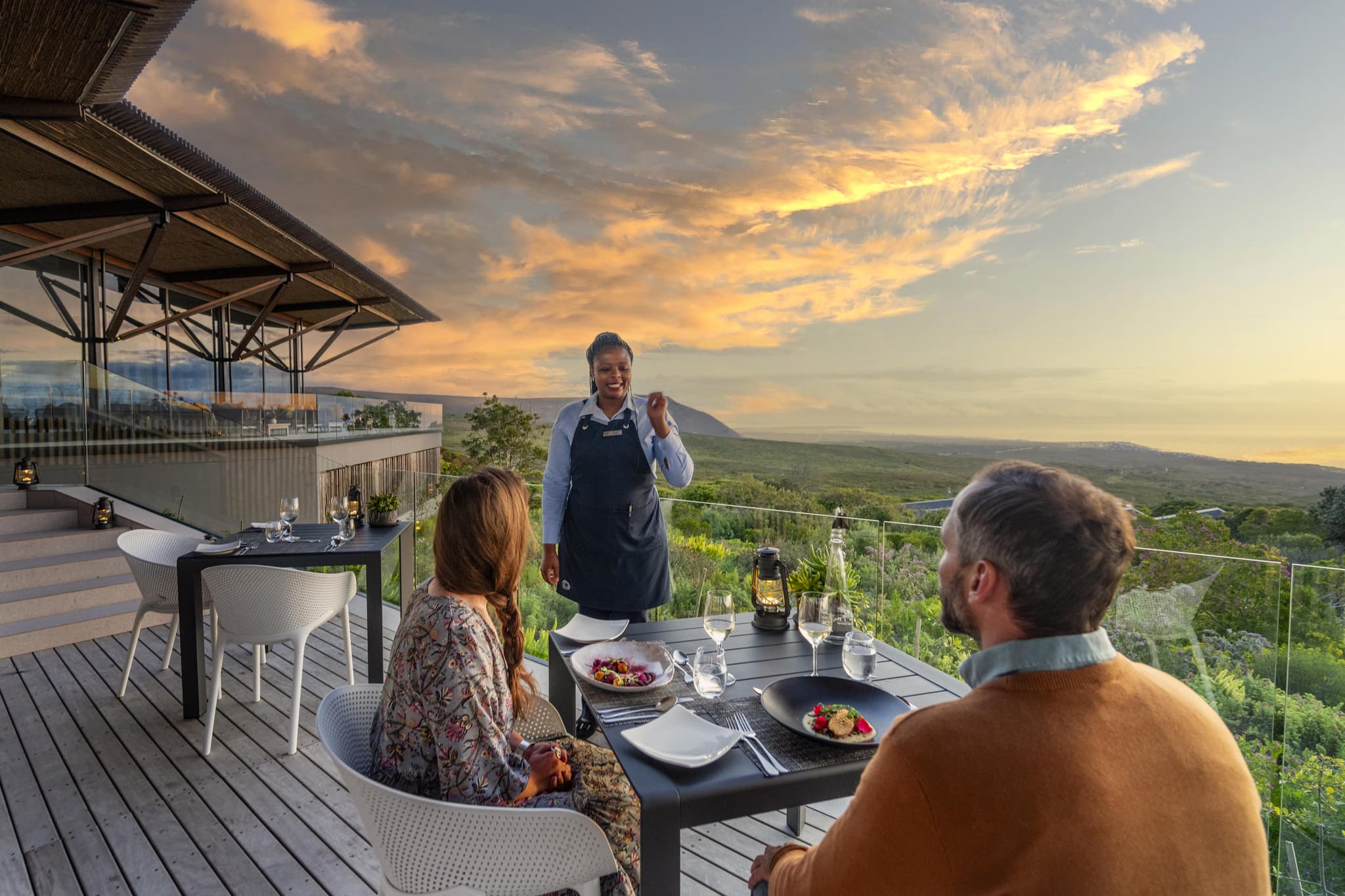 Couple being served dinner on a luxury lodge terrace overlooking mountains at sunset