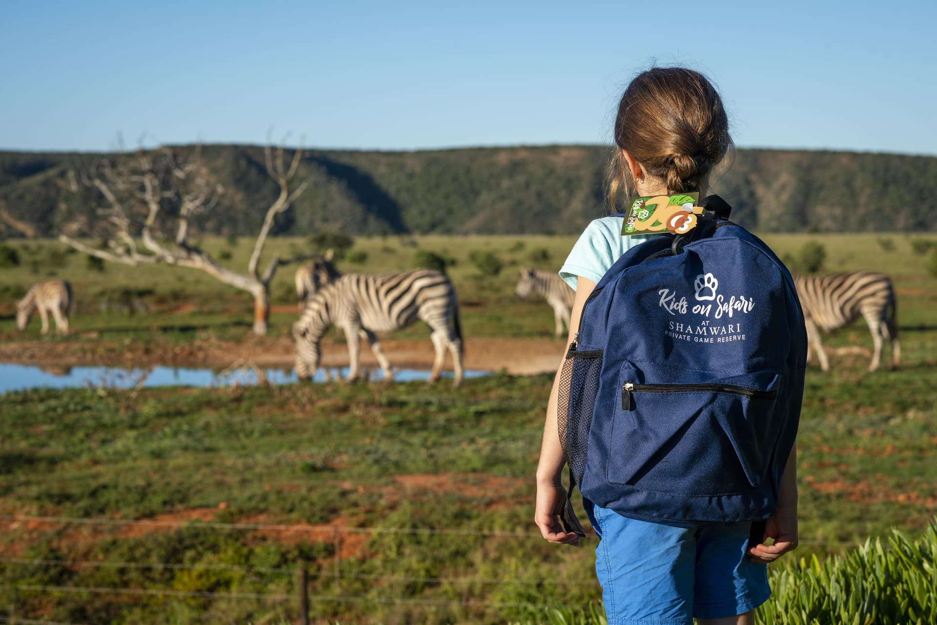 Young girl with Shamwari Kids on Safari backpack watching zebras at a waterhole